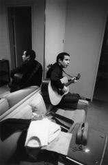 Paul Simon sitting on arm of couch holding a guitar