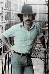 Young Paul Simon with mustache and wearing a hat standing on a fire escape