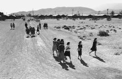 people walking down a barren dirt road with barracks visible in the distant background