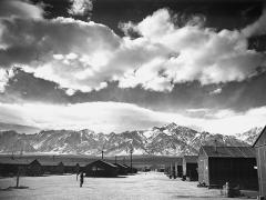 a single person stands among wooden barracks with dramatic clouds in sky above and mountains visible in distance