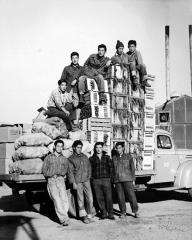 crates and cloth bags piled high in the back of a truck with men sitting on top of the crates and standing next to the truck