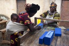 a woman carrying a baby on her back with another woman at a work table with blue blocks on it