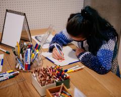 Photo of a child drawing at a large, wooden desk. The desk is filled with cups of pencils, crayons, and markers.