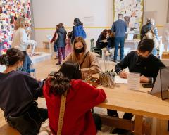 Photo of a gallery filled with people. Some are looking at a quilt on the far wall, some are walking through the space, and four are seated at a table in the foreground writing and drawing together.