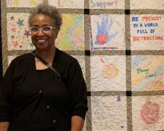Image of the artist Pam Johnson standing in front of a community quilt of different sourced squares. She is wearing all black and smiling at the camera.