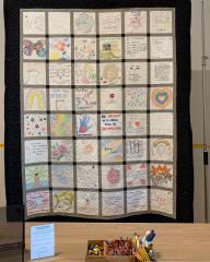 A front-facing photo of a large quilt hanging from a tall structure behind a table. The quilt is made up of 48 white squares that have been decorated by different individuals.