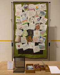 A front-facing photo of a large quilt handing from a tall structure behind a table. The quilt is made up of many, overlapping white squares that have been decorated by different individuals.