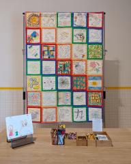 Photo of a large quilt hanging from a tall structure behind a table. The quilt is made of 35 white squares that have been decorated by different individuals. Colored lines mimicking a rainbow attach each square.