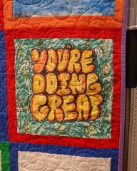 Detail photo of a quilt showing a decorated square with the words "You're Doing Great" drawn on it in yellow and orange.