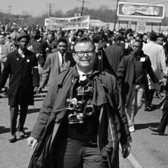 Black and white photo of Bob Adelman walking on a street in front of a large group of people. His cameras are hanging around his neck.