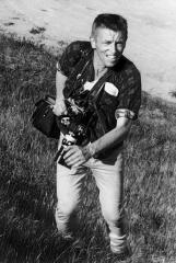 Black and white photo of George Ballis in a large, grass field holding his camera and looking at something to his left.