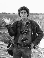 Black and white photo of Bob Fitch facing the camera and waving. He is standing before a large, grass field and has two cameras around his neck.