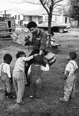 Black and white photo of Bob Fletcher standing in front of a house showing a group of four small children his camera.