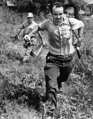 Black and white photo of Matt Herron running through a large, grass field with a camera in each hand. Behind him is a law enforcement officer running with a batton.