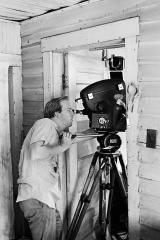 Black and white photo of David Prince standing in a doorway looking through the viewfinder of a film camera set on a tripod.