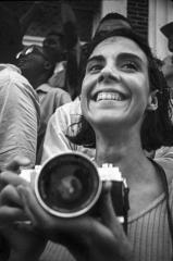 Black and white close up photo of Maria Varela smiling and holding her camera. Behind her is a group of people engaged in a chant.