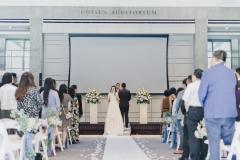 Photo of a wedding ceremony looking down the aisle towards the stage where guests, bride, and groom are standing with their backs to the camera.