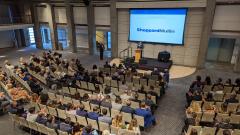 A large auditorium filled with people watching a presentation on stage with a large screen.