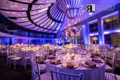 The ballroom set up for an evening event with dining tables, colored lights, and large floral bouquets.