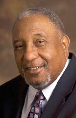 A headshot of an African American man in a dark suit with a muted colored tie with squares on it. He has black and grey short hair and a goatee.