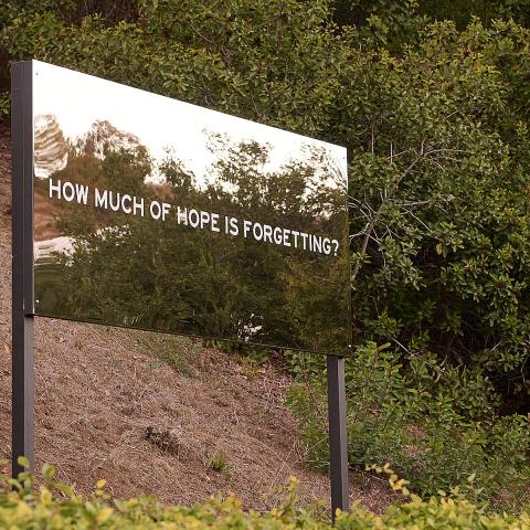 A large reflective billboard on a hillside that says, "How much hope is forgetting?". An outdoor artwork by Chloe Bass.