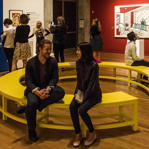 A man and women seated on a yellow circular bench in the middle of a gallery with people walking about