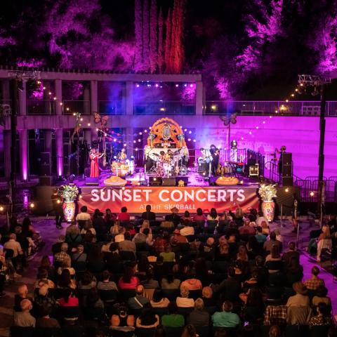 Wide view of a nighttime concert with performers playing and a large seated audience watching. A big stage banner reads Sunset Concerts at the Skirball.