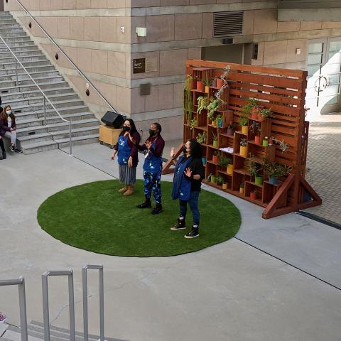 Three educator performing a story in front of an audience kids and adults in an outdoor theater. There are standing on green grass-like circular matt with a wooden backdrop with colorful potted plants.