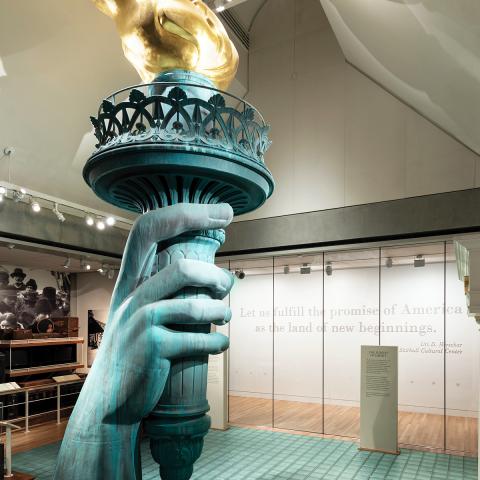 Photo inside the exhibition Vision and values showing a replica of the statues of liberty hand holding the torch among a large photo mural of immigrants on Ellis Island