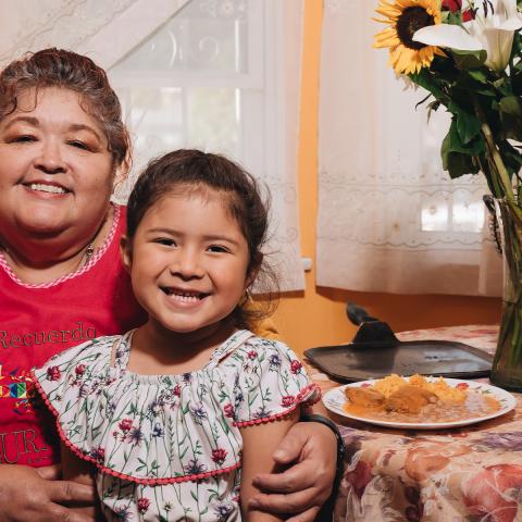 Photo of a woman and girl sitting together at a colorfully decorated table. They are both smiling at the camera. On the table are plates of food, a vase of colorful flowers, and a piñata.