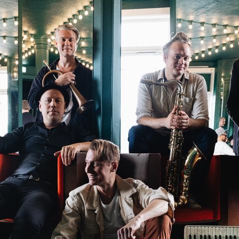 Photo of the six member band sitting in a mirrored room. Each member is holding their instrument and smiling at the camera.