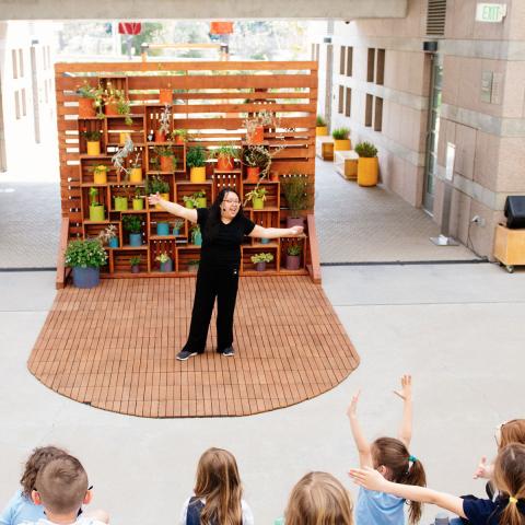 Educator in performing a story with arms stretched wide while children are watching from outdoor amphitheater seats. There is a wooden planked backdrop with color potted plants.