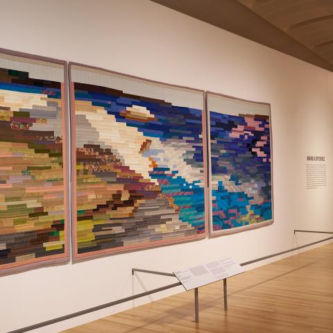 Image of a gallery space. Colorful quilts are seen hanging on white walls. Closest to the frame is a triptych quilt displaying a coastline and ocean made up of colorful bands of fabric.