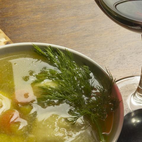 Overhead photo of a bowl filled with matzo ball soup. Next to the bowl is an unleavened cracker, glass of red wine, and spoon.