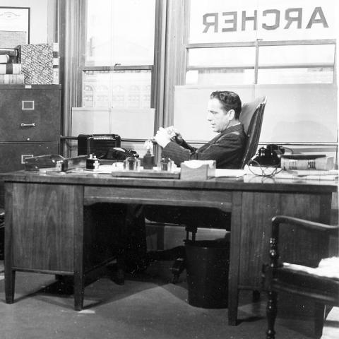 Black and white image of two men sitting at desks in an office conversing. The name "Archer" is painted on the windows behind them.