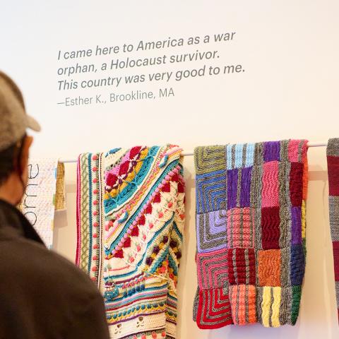 Photo of a man in a baseball cap facing a white wall with many colorful blankets displayed.