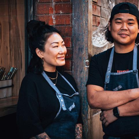 Two people standing in front of a brick building with cutlery out in pails. They are wearing black shirts and blue aprons smiling.