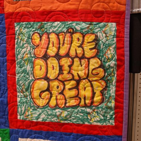Detail photo of a quilt showing a decorated square with the words "You're Doing Great" drawn on it in yellow and orange.