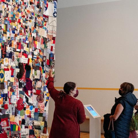 A women is pointing at a large community quilt hanging patched together with many different pieces. A family is standing next to her observing.