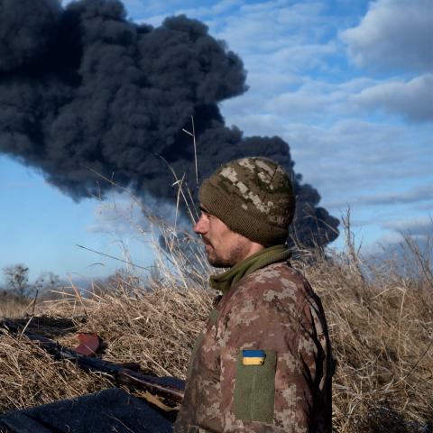Photo of a Ukrainian soldier in a field with his dog. Both are looking at something off camera to the left. Behind the man is a large plume of black smoke filling the skyline.