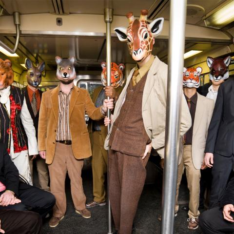 Photograph of the 11 band members sitting and standing in a subway car. They are all wearing suits and animals masks and looking at the camera.