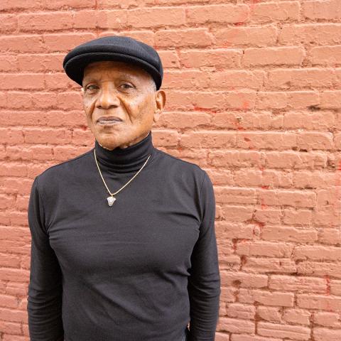 The artist dressed in all black and standing in front of a red brick wall looking off camera.