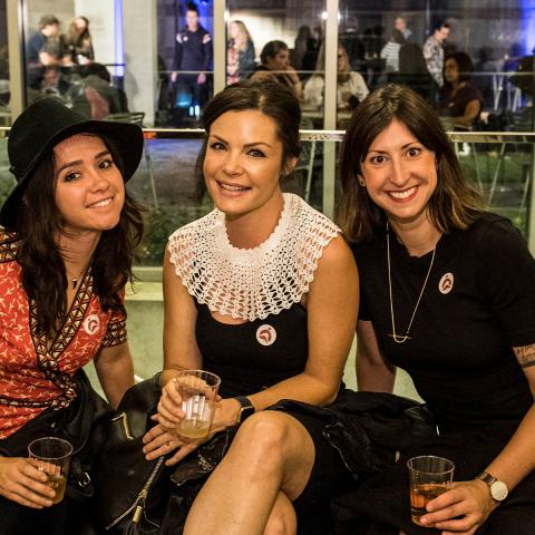 Three women sit together on a bench smiling at the camera. Behind them a party can be seen through a wall of windows.