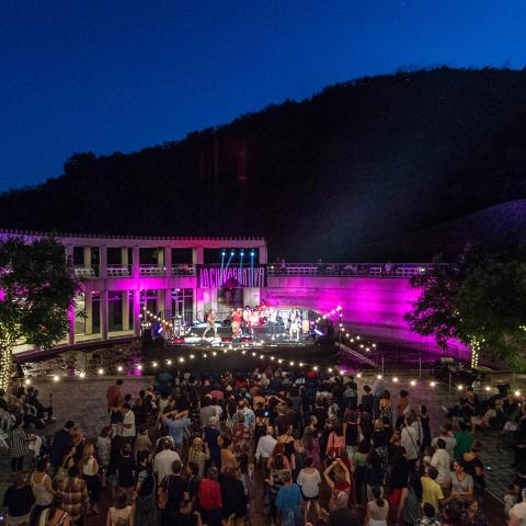 Photo of an outdoor concert at the Skirball. A large group of people are in the courtyard dancing to a band on stage. Lights are seen in the trees, strung across the courtyard, and from the stage.