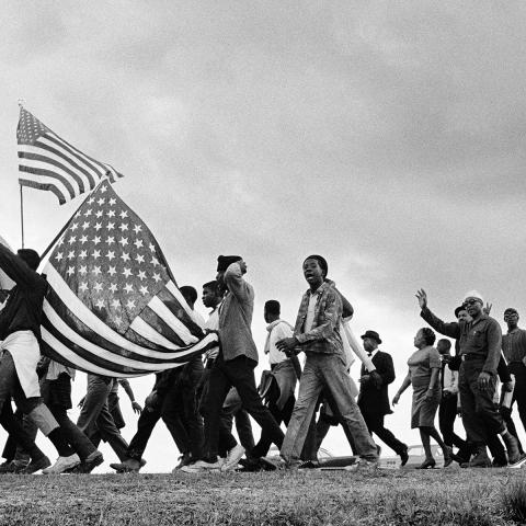 Black and white photo of a large group of people walking on a dirt road from right to left. Two people to the left side of the image are holding American flags.