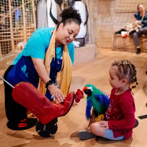 An educator kneels by a young child engaging with two animal puppets in the Ark