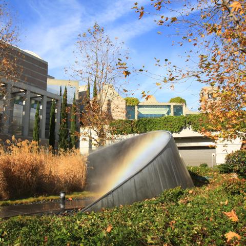 A metal sculpture that shaped in an arc emits mist and creates a rainbow. The sculpture sits in an green arroyo outside of a 3 story building.