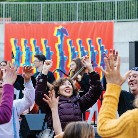 A group of people dancing outside in front of a large banner of a hanukkiah