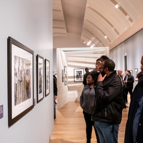 Many people standing in the exhibition This Light of Ours looking at a wall of black and white photographs