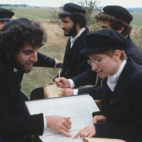 Still from the movie showing Mandy Patinkin and Barbara Streisand sitting with other men in the back of a truck reading religious text